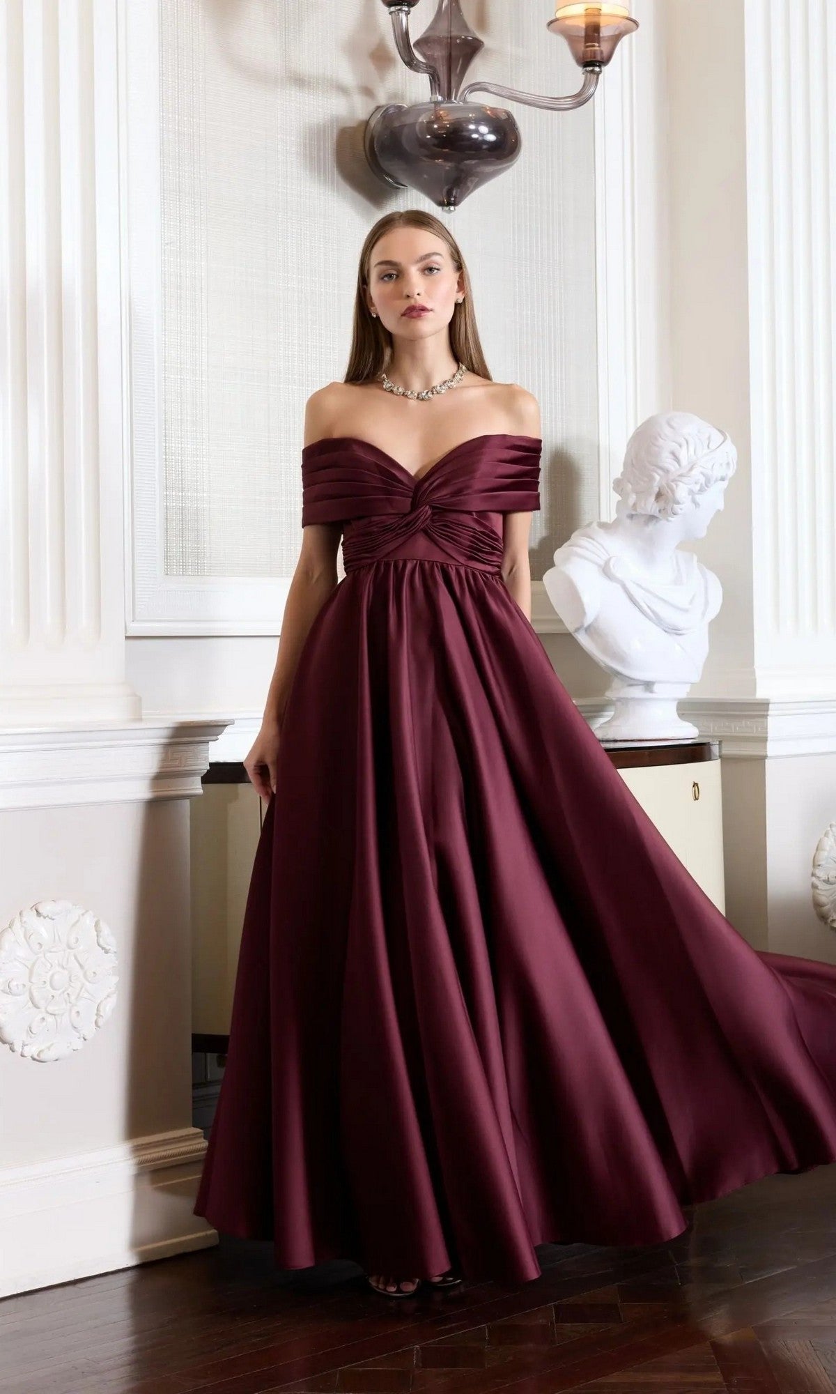 A woman in the wine-colored Mac Duggal 56188 long formal dress poses indoors by a white classical bust and ornate wall, highlighting the elegance of her off-the-shoulder evening gown.